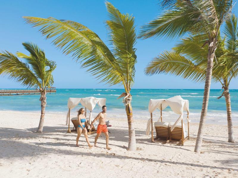 Strand mit Palmen, blauem Himmel, Meer und entspannten Menschen in sommerlicher Kleidung.