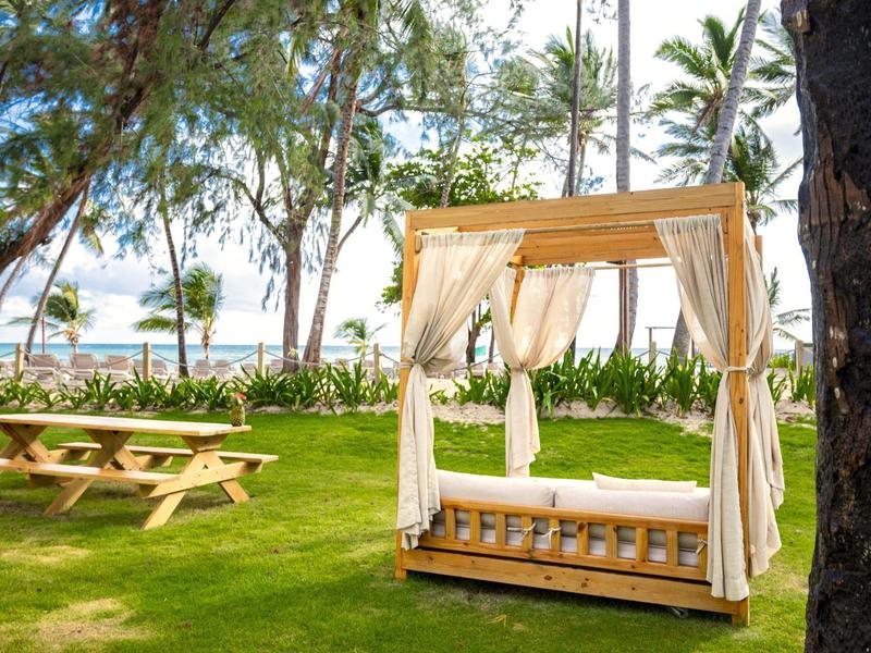Ein gemütliches, überdachtes Strandbett mit weißen Vorhängen steht auf grünem Rasen neben einem Holztisch.