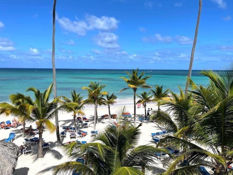 Strand mit Palmen, weißen Sand und blauem Meer unter hellem Himmel mit vereinzelten Wolken.