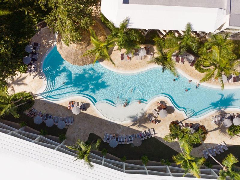 Großer, geschwungener Pool mit blauen Liegestühlen, grünen Palmen und Sonnenschirmen umgeben.