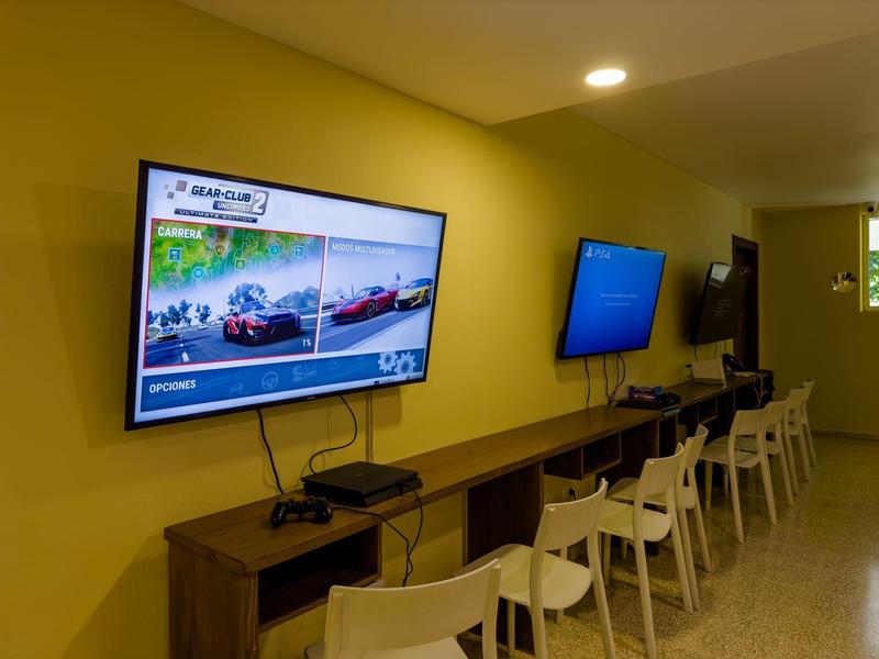 Mehrere weiße Stühle vor einer langen Holztheke mit zwei Wandbildschirmen, gelbe Wand im Hintergrund.