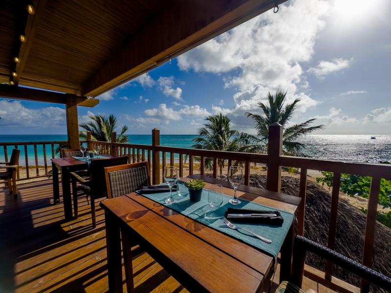 Holzterrasse mit gedecktem Tisch und Blick auf Palmen, Strand und blauen Himmel am Meer.