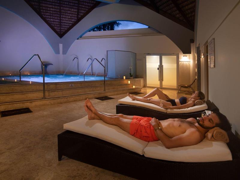 Ein Schwimmbad mit beleuchtetem Wasser und Menschen, die auf Liegen entspannen, helle Wände und Dachstuhl sichtbar.