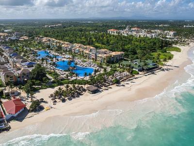 Vista aérea de una playa con instalaciones hoteleras, piscina y palmeras junto al mar.
