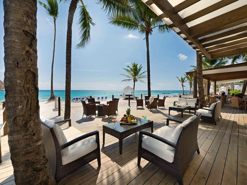 Área de asientos abierta con sillones blancos con vista a la playa y al mar bajo palmeras.