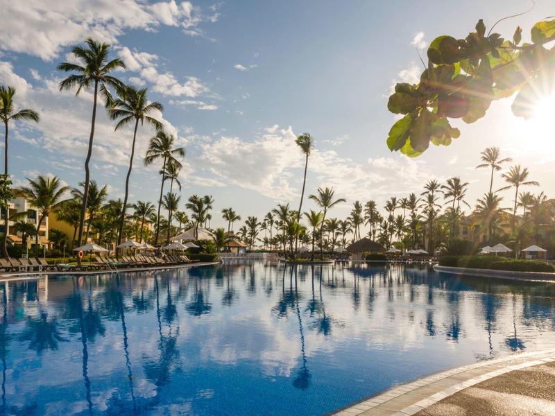 Gran piscina con palmeras y luz solar en un resort junto al mar.