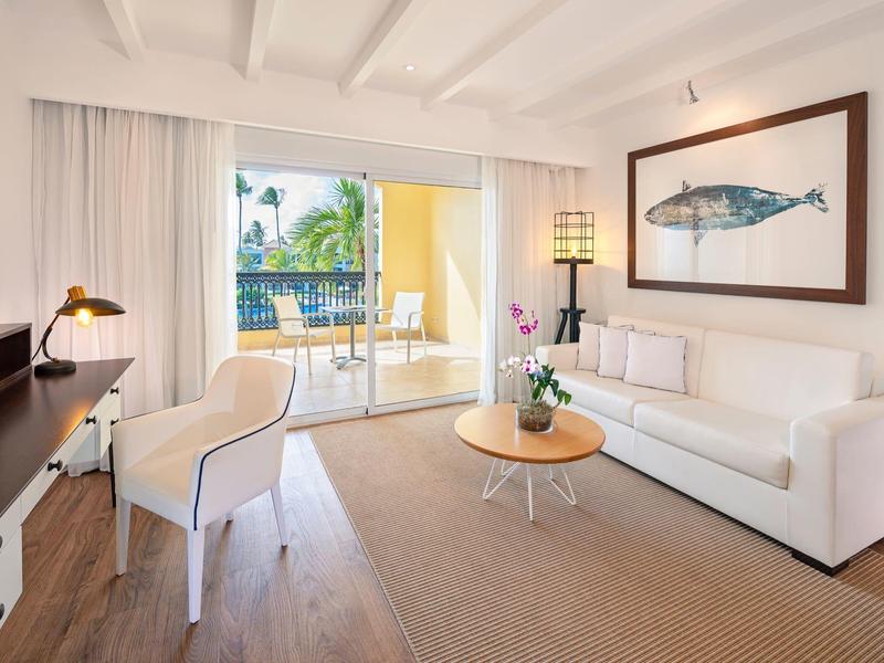 Sala luminosa con sofá, escritorio y acceso a un balcón con plantas.