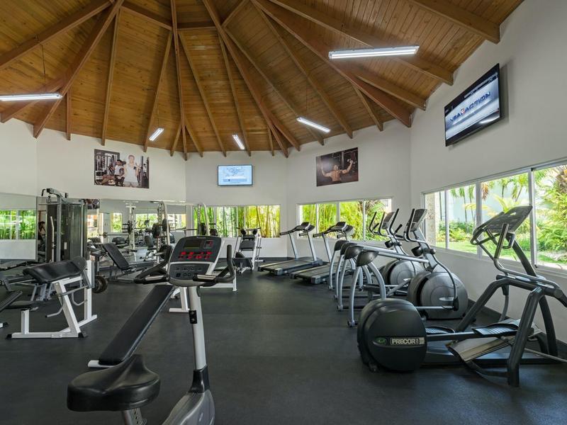 Gimnasio moderno con equipos y techo de madera, ventanas grandes con vista al verde.