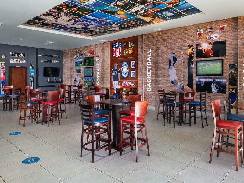 Interior de un bar deportivo con mesas de madera, sillas rojas y decoraciones deportivas en las paredes.