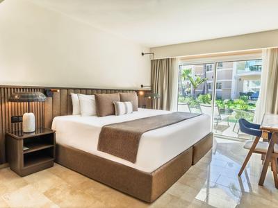 Camera d'hotel luminosa con letto matrimoniale e accesso a una terrazza con vista sul giardino e piscina.