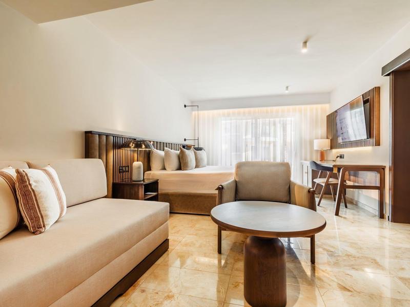 Moderní hotelový pokoj s pohovkou, křeslem, stolem a televizí na zdi.