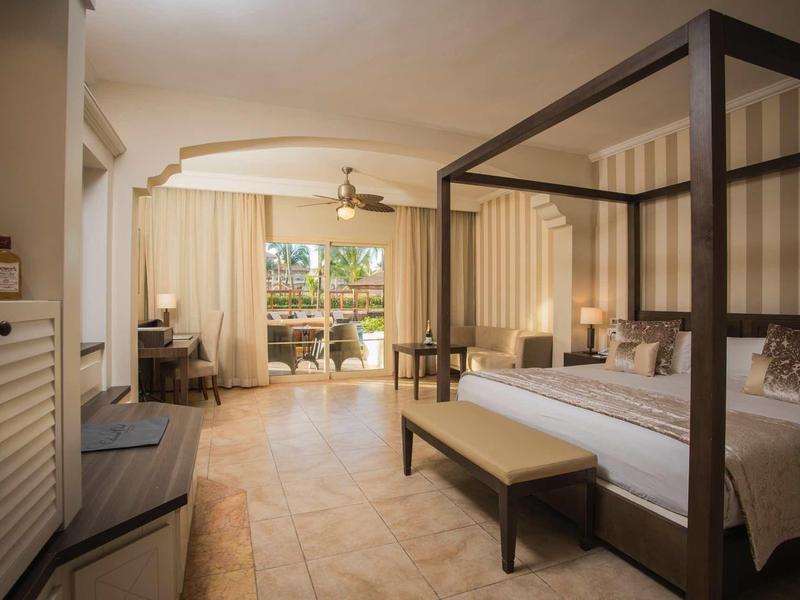 Bright hotel room with canopy bed, bench, and balcony overlooking garden.