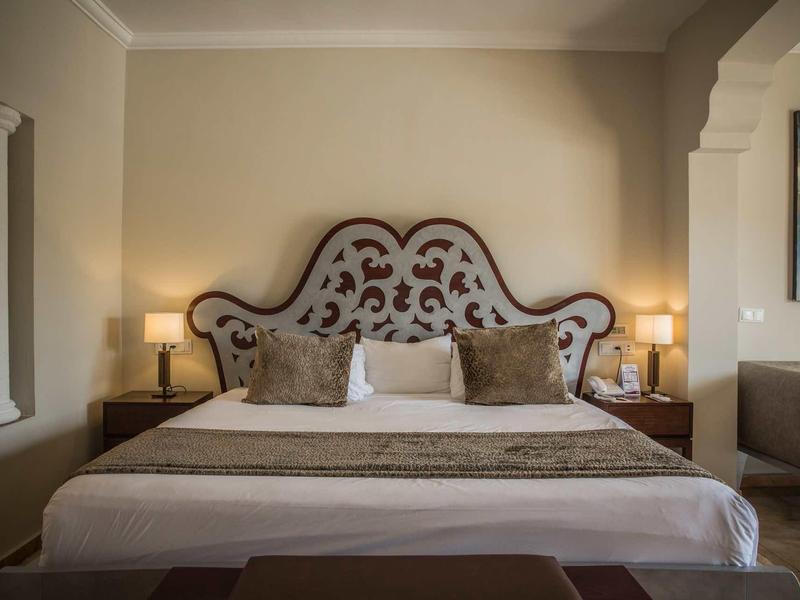 Elegant hotel room with large bed, decorative headboard, and warm lighting.