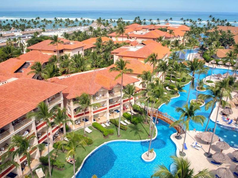 Large resort with red roofs, expansive pool, and sea view under clear weather.