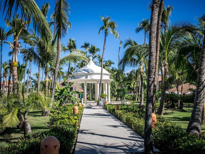 A manicured garden path with palm trees leads to a white pavilion structure under a blue sky.