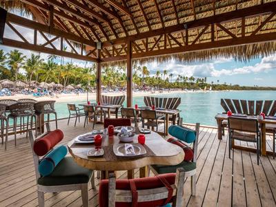 Open-air restaurant with wooden floor and sea view, wooden tables and chairs under a thatched roof.