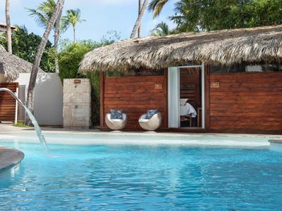 Bungalow mit Strohdach neben blauem Pool, Palmen im Hintergrund und Wasserspeier im Poolrand.