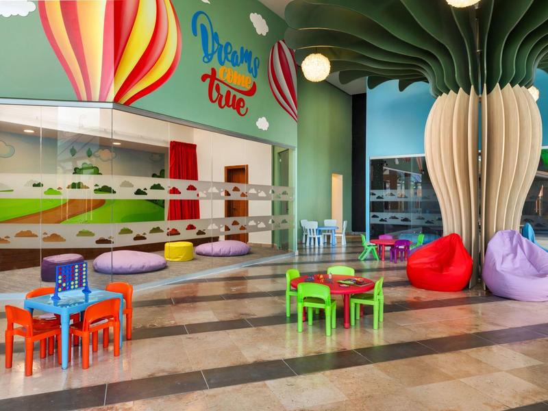 Aire de jeux colorée pour enfants avec petites tables, chaises et grandes plantes décoratives dans le hall de l'hôtel.