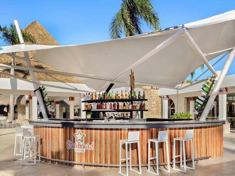 Bar ouverte avec bois et tabourets blancs sous des parasols en plein air.