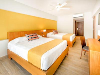 Helles Hotelzimmer mit zwei Betten, Holzmöbeln, gelber Wand und Deckenventilator.