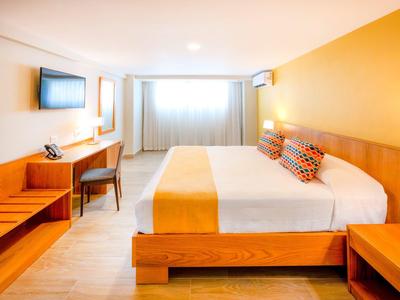 Helles Hotelzimmer mit großem Bett, Holzmöbeln und gelb-orangen Akzenten an Wand und Bettwäsche.