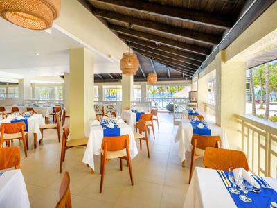 Geräumiges Restaurant mit weißen Tischdecken, orangen Stühlen und Holzdachbalken, Blick auf Palmen.