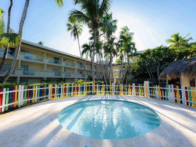 Runder Pool mit klarem Wasser, umgeben von buntem Zaun und hohen Palmen vor Hotelgebäude.