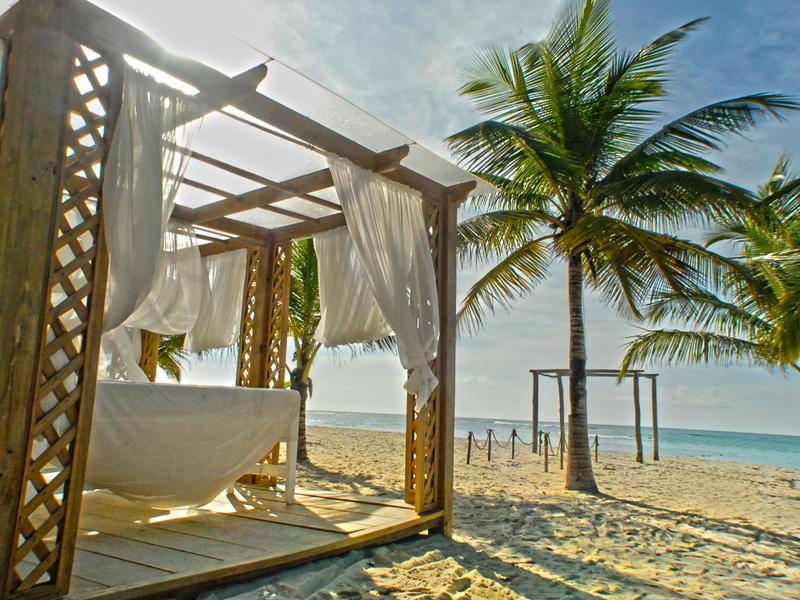 Freestanding bathtub under wooden pavilion on the beach with palm trees and sea view.