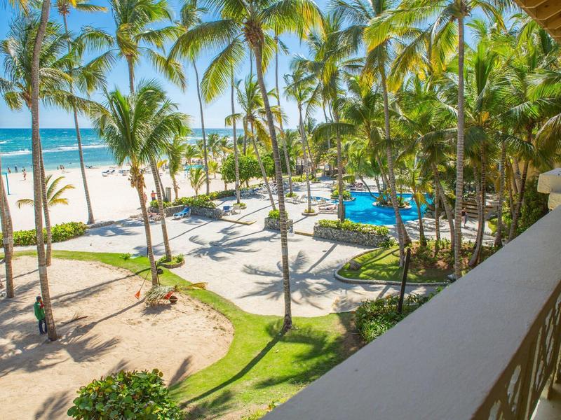 View from a balcony overlooking a tropical beach with palm trees and a pool at the hotel resort.
