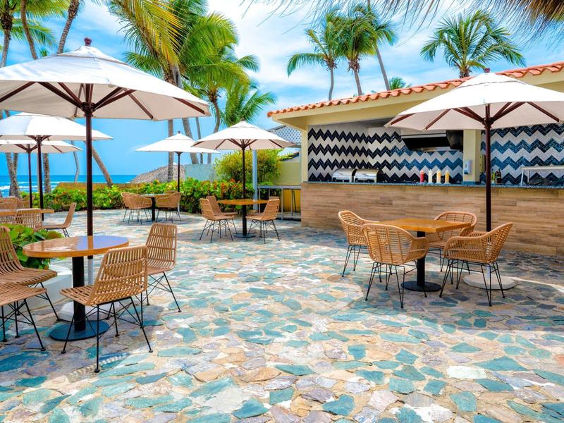 Outdoor restaurant with umbrellas, tables, and chairs against a tropical seaside backdrop.
