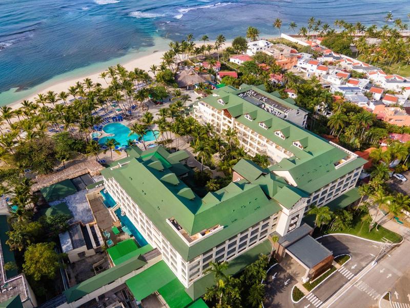 Aerial view of a resort with green roofs, pool, and palm-lined beach by the sea.