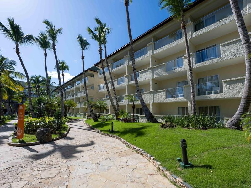 Modern hotel complex with balconies, palm trees, and manicured garden on a sunny day.