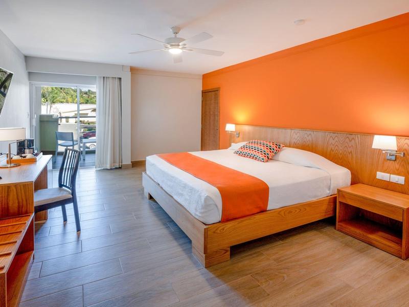Modern hotel room with double bed, orange accent wall, and desk with chair.