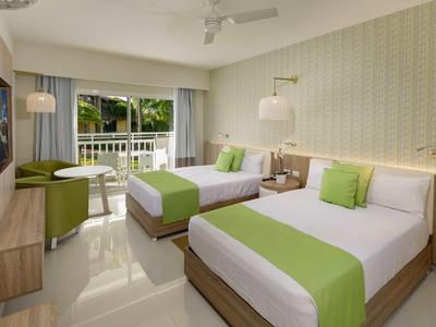 Helles Hotelzimmer mit zwei Betten, grün-weißer Bettwäsche und Balkonblick auf Pflanzen.