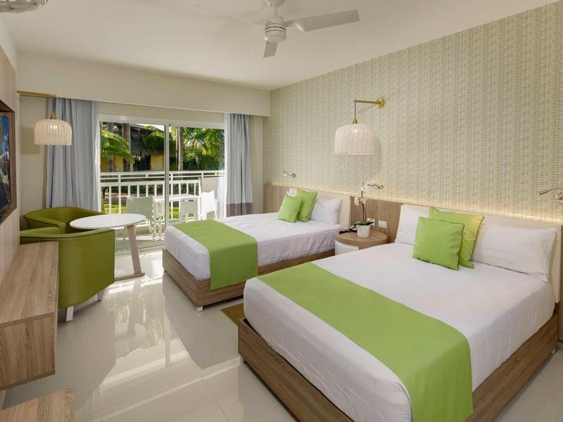 Helles Hotelzimmer mit zwei Betten, grün-weißer Bettwäsche und Balkonblick auf Pflanzen.