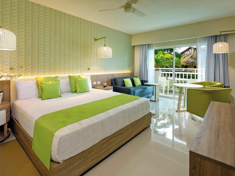 Modern eingerichtetes Hotelzimmer mit großem Bett und Balkon mit Blick auf tropische Pflanzen.