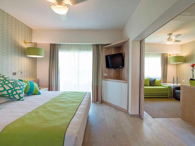 Modernes Hotelzimmer mit Doppelbett, grünem Bettläufer und angrenzendem Wohnzimmer mit Sofa.