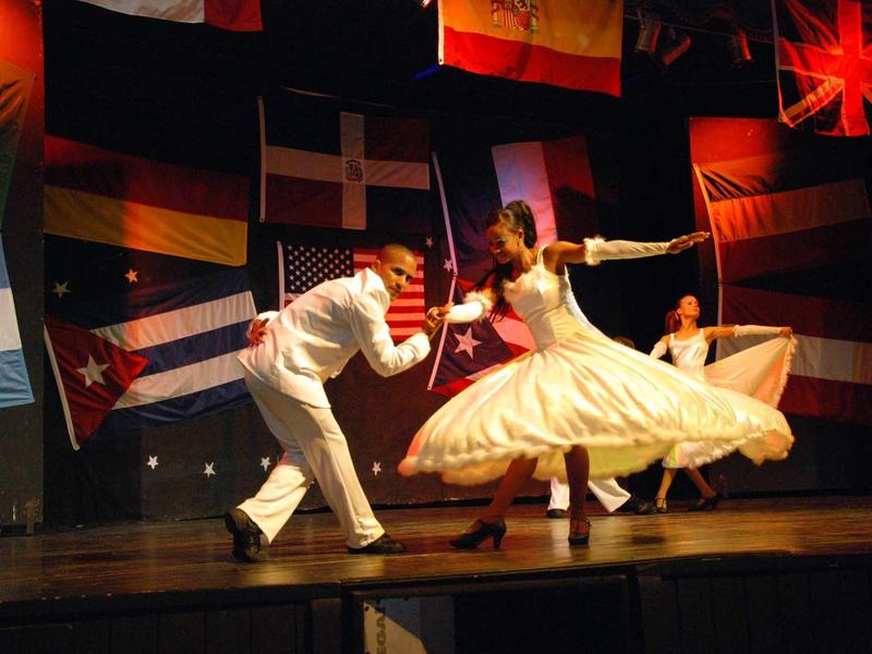 Tänzer im weißen Outfit wirbeln auf Bühne mit vielen bunten Länderflaggen im Hintergrund.