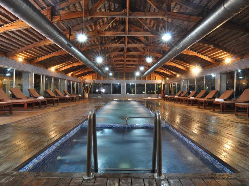 Innenraum eines Hallenbads mit Holzdecke, Holzboden, Wasserspiegel und Reihen von Liegestühlen.