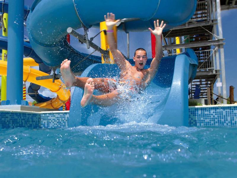 Zwei Personen rutschen eine blaue Wasserrutsche hinunter, Wasser spritzt um sie herum.