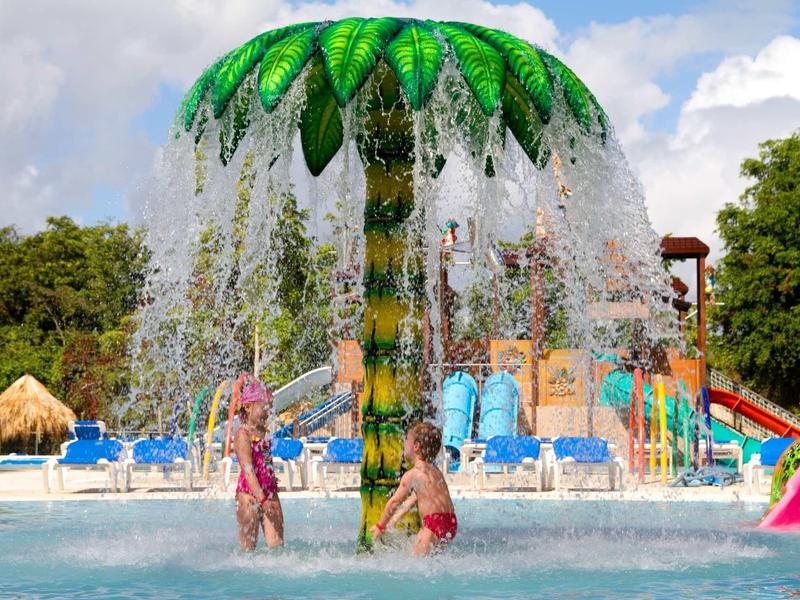 Wasserspiel im Spaßbad mit zwei Kindern unter einem grünen Pilzregen und blauen Liegen im Hintergrund.