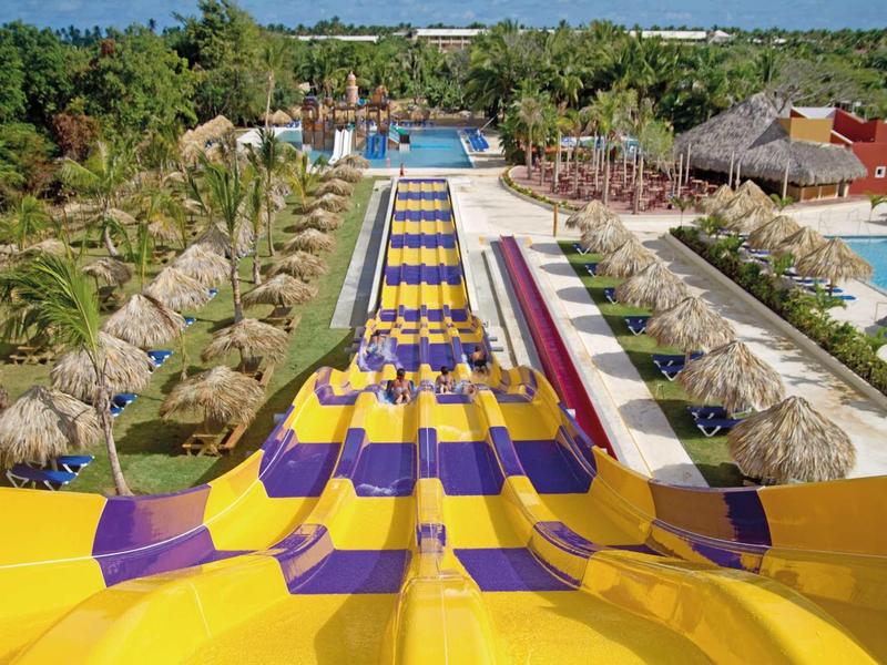 Bunte Wasserrutschen in Gelb und Lila führen hinab zu einer Poollandschaft mit Palmen und Sonnenschirmen.