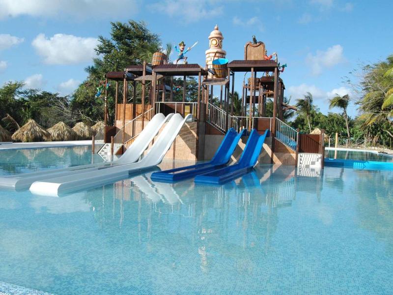 Großes Schwimmbecken mit drei blauen Rutschen und Palmen im Hintergrund unter blauem Himmel.