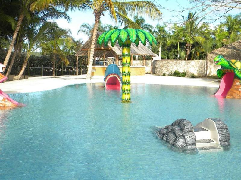 Kinderpool mit bunten Rutschen und Palmen-Sprinkler in klarem, blauem Wasser und Sonnenschein.