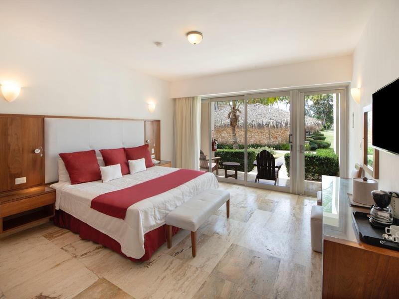 Bright hotel room with large bed, red accents, and terrace view of garden.