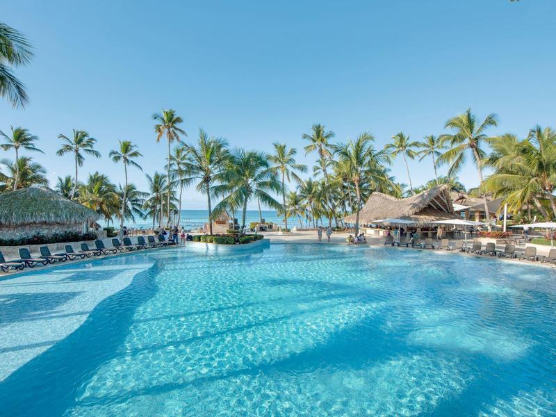 Large swimming pool with lounge chairs and palm trees under clear sky by the sea.