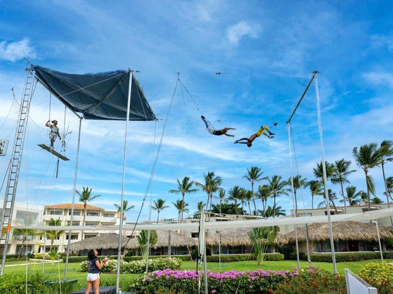 People trapezing over a tropical resort with palm trees and blue sky.