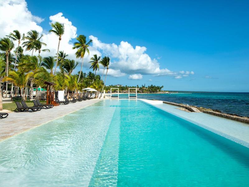 Infinity pool by beach with palm trees and blue sky on a sunny day.