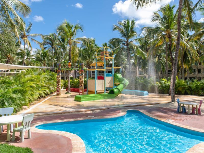 Outdoor pool with children's playground and palm trees at tropical resort.