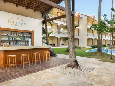 Área de bar al aire libre con taburetes de madera junto a piscina y edificio hotelero en entorno tropical.
